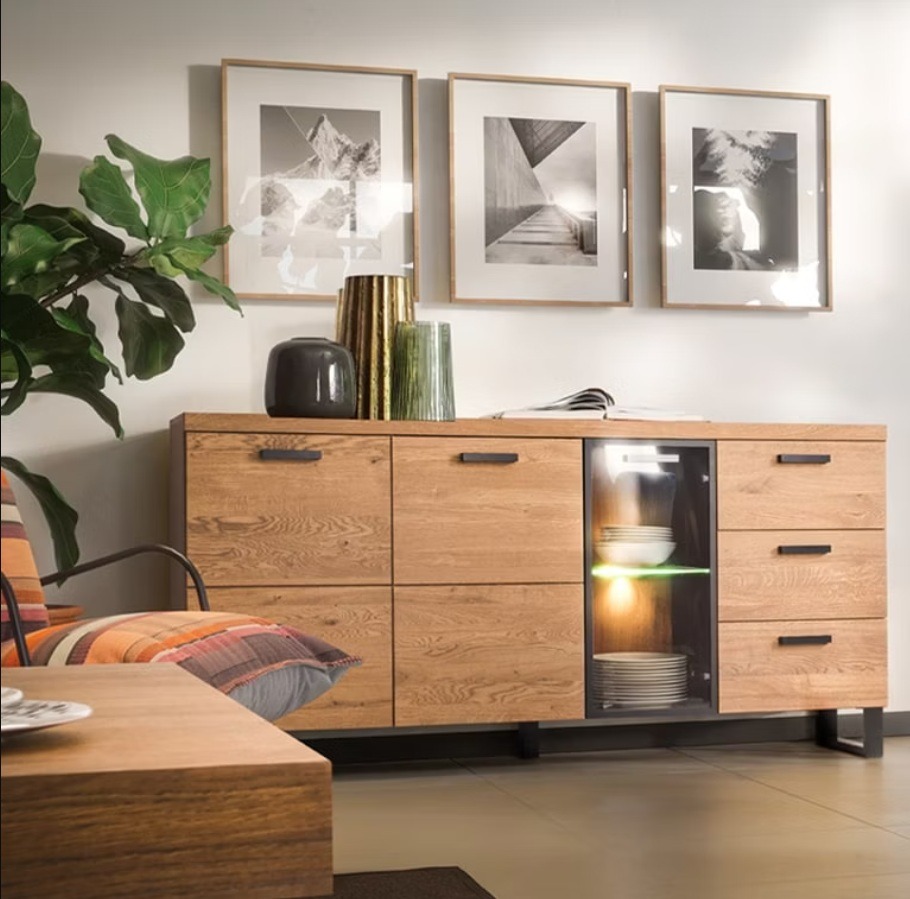 Drewniana komoda z przeszkloną witryną, oświetlona od wewnątrz, z trzema obrazami w ramkach na ścianie i rośliną doniczkową obok. Nowoczesny design.