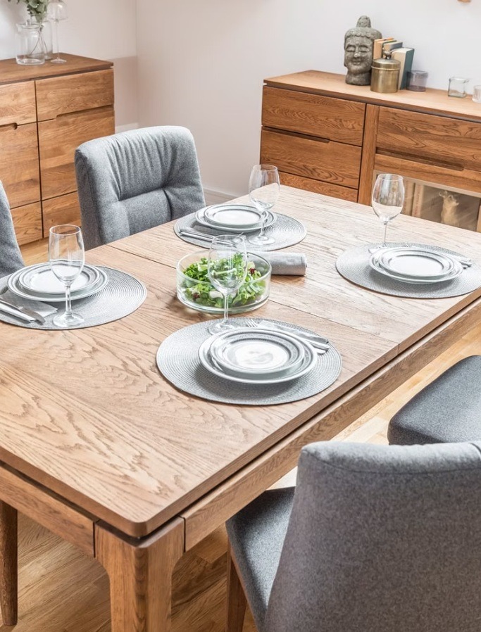 Drewniany stół z szarymi krzesłami, nakryty do posiłku z sałatką w szklanej misce. W tle komoda z drewnianymi frontami i dekoracyjna figurka Buddy.