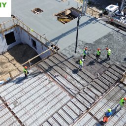 PROJEKTY DOLNY SP. Z O. O. - Wysoka perspektywa na budowę stropu: ekipa w kaskach nadzoruje wylewanie betonu z pompy. Widoczne zbrojenia i pustaki szalunkowe. Część stropu już zalana.