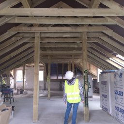 PROJEKTY DOLNY SP. Z O. O. - Wnętrze w trakcie budowy z drewnianą konstrukcją dachu, widok z perspektywy osoby w kasku i kamizelce odblaskowej. W tle okna i materiały budowlane.