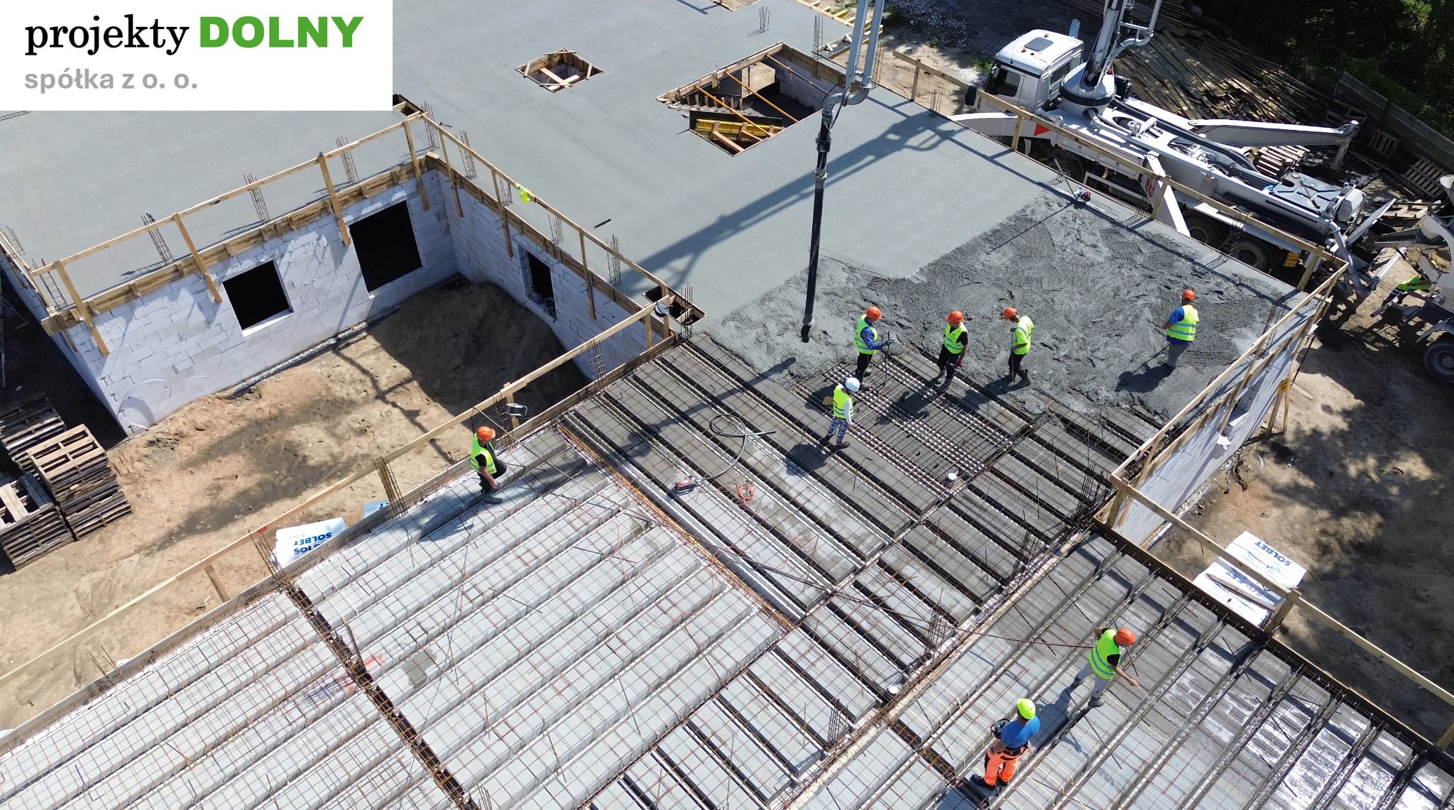 Wysoka perspektywa na budowę stropu: ekipa w kaskach nadzoruje wylewanie betonu z pompy. Widoczne zbrojenia i pustaki szalunkowe. Część stropu już zalana.