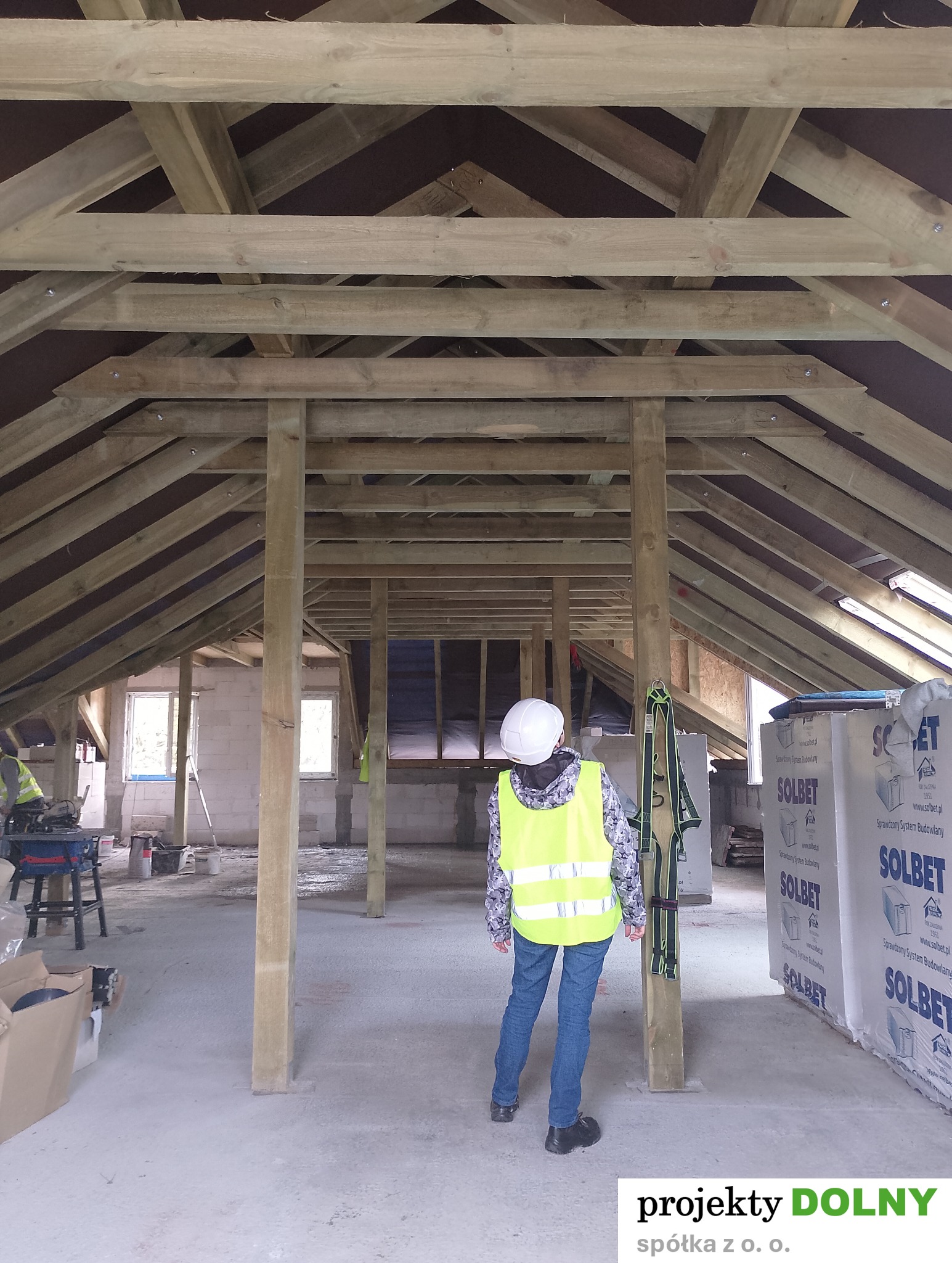 Wnętrze w trakcie budowy z drewnianą konstrukcją dachu, widok z perspektywy osoby w kasku i kamizelce odblaskowej. W tle okna i materiały budowlane.