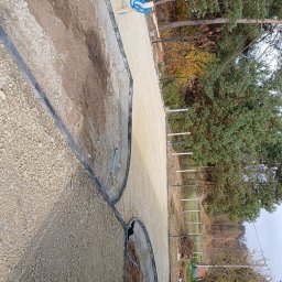 Fhu - Przygotowany teren pod ścieżkę z obrzeżami, podsypany żwirem. Widoczne narzędzia i pracownicy. W tle drzewa i budynek.