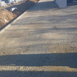 Fhu - Utwardzona droga dojazdowa z krawężnikiem, w tle budynki mieszkalne i koparka. Widok z perspektywy osoby stojącej na drodze, w słoneczny dzień.