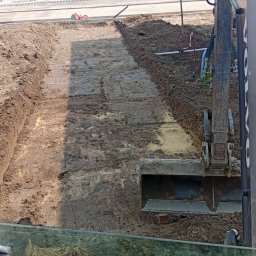 Fhu - Głęboki wykop w ziemi, widoczny z kabiny koparki Volvo. W tle budynek z szarymi bramami garażowymi i inny sprzęt budowlany. Etap prac ziemnych.