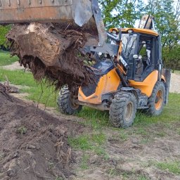 Fhu - Żółta koparko-ładowarka MST podnosi bryłę ziemi z korzeniami na tle zieleni. Prace ziemne w toku, widoczne ślady opon na gruncie.