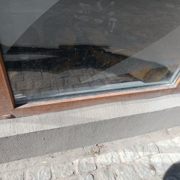 Jaroslav Dvoran - Mycie Okien Bydgoszcz - Brudna szyba okienna z drewnianą ramą, widoczne zacieki i kurz. W tle zarys otoczenia. Dolna część okna przylega do szarego muru i kamiennej kostki brukowej.