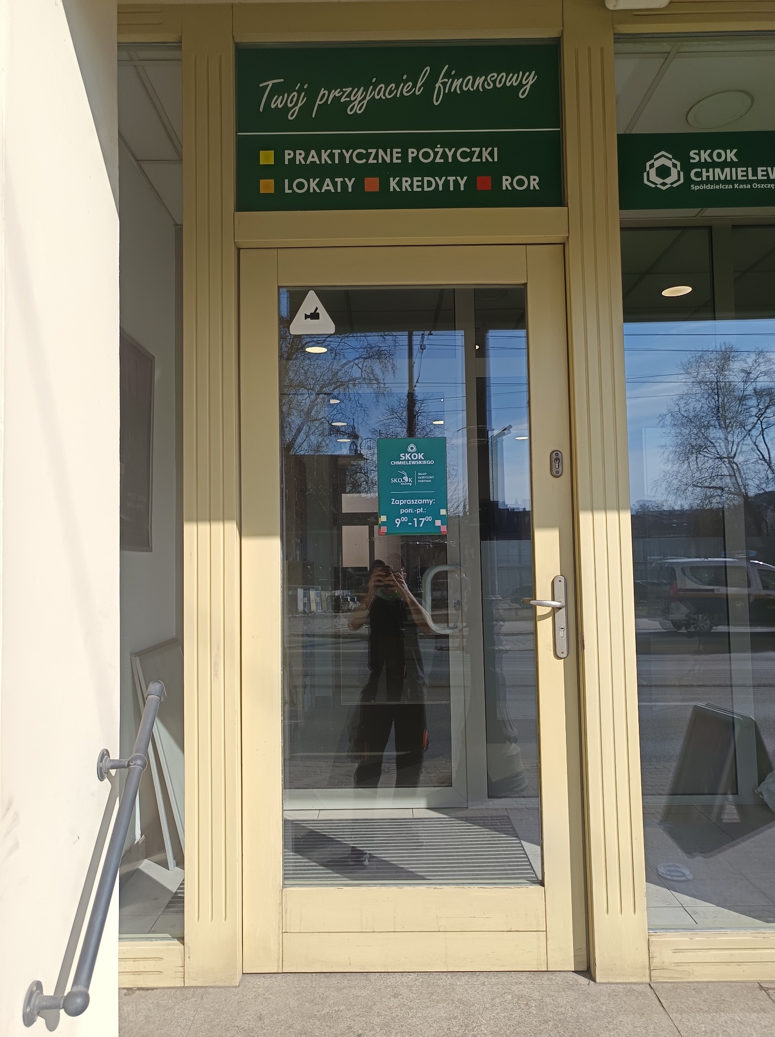 Przeszklone drzwi wejściowe do placówki SKOK Chmielewskiego z widocznym plakatem informacyjnym i odbiciem osoby. Jasna elewacja budynku w słoneczny dzień.
