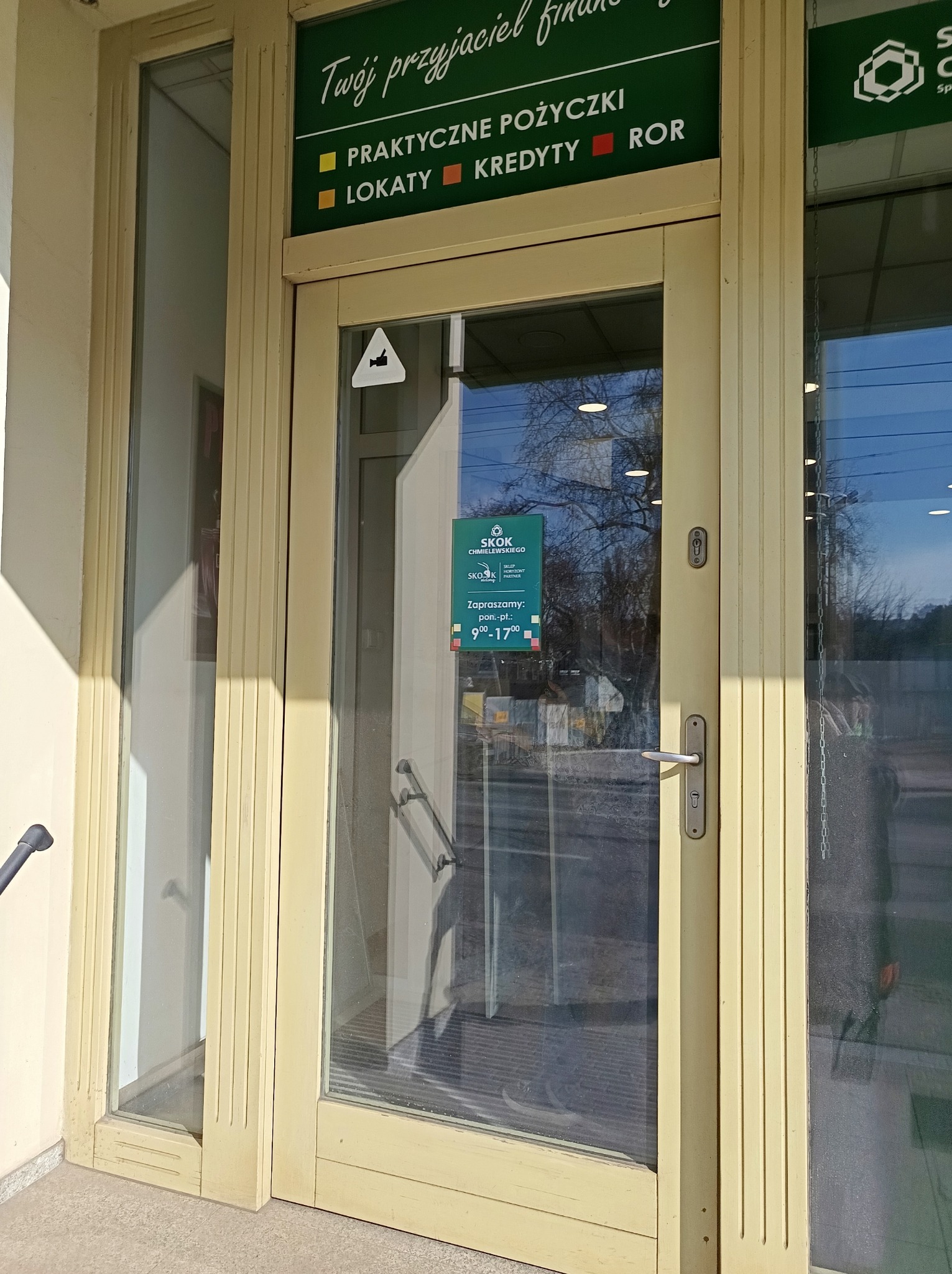 Jasne, przeszklone drzwi wejściowe do placówki finansowej z widocznym plakatem informacyjnym i odbiciem otoczenia. Widoczne zabrudzenia na dolnej części szyby.