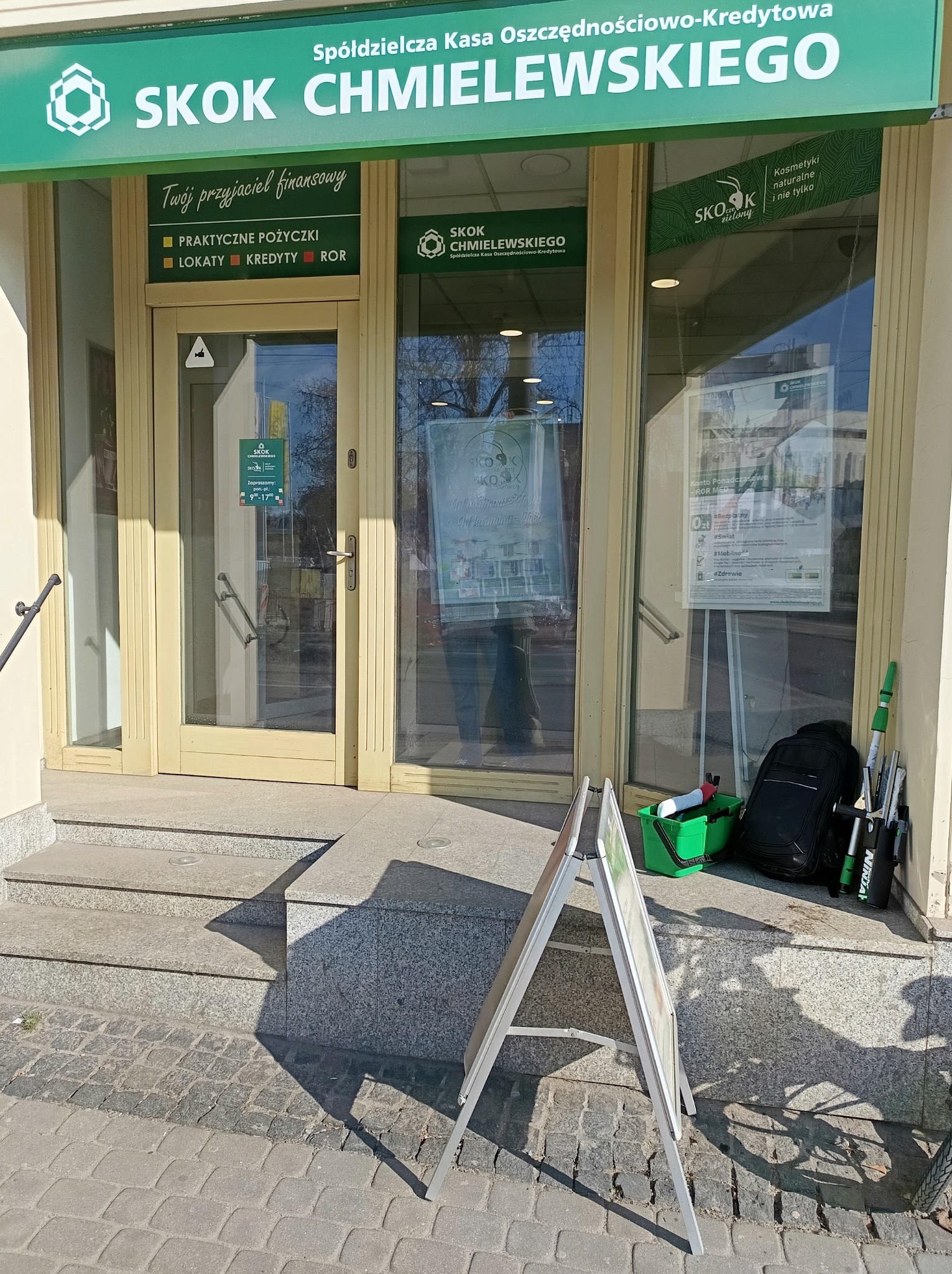 Sprzęt do mycia okien (wiaderko, myjki, kij teleskopowy) ustawiony przed wejściem do placówki SKOK Chmielewskiego. Na pierwszym planie tablica reklamowa.