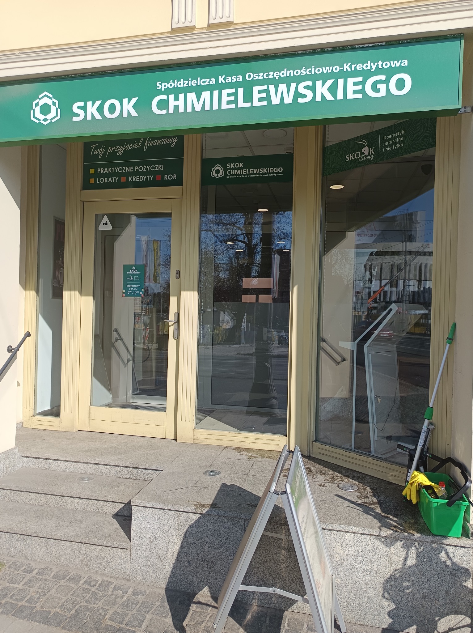 Wejście do placówki SKOK Chmielewskiego z wiadrem i mopem do czyszczenia okien. Widoczny szyld i drzwi z szybami. Na chodniku stoi tablica reklamowa.
