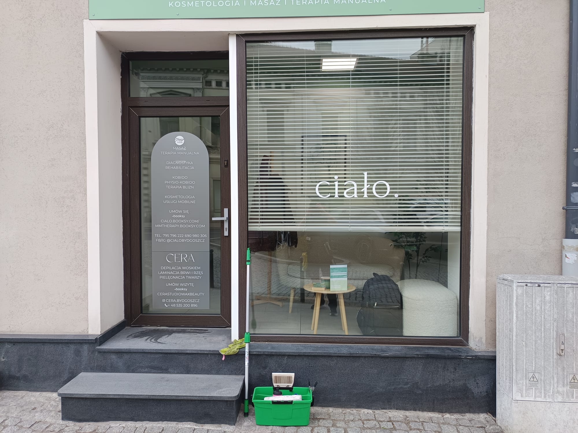 Mycie okien salonu kosmetycznego: zielony pojemnik z narzędziami do mycia, mop oparty o szybę z napisem 'ciało.'. Widoczne odbicie wnętrza.