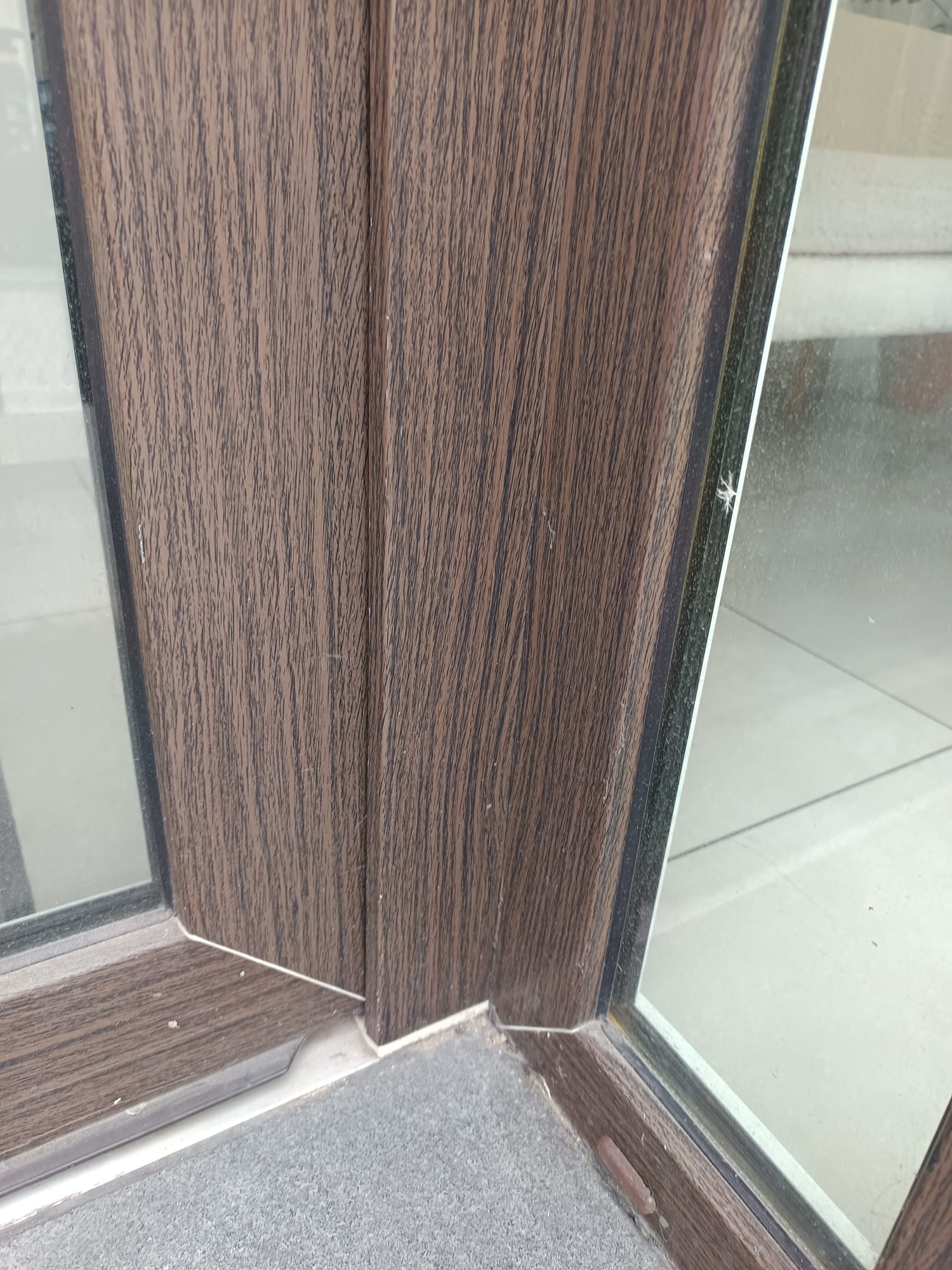 Brudne okno z drewnopodobną ramą, widoczne zabrudzenia na szybie i ramie, szara podłoga. Detal narożnika okna przed czyszczeniem.