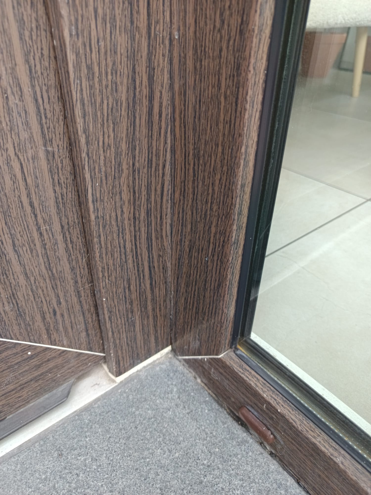 Fragment okna z brązową ramą o strukturze drewna, widoczny narożnik i dolna krawędź, szara podłoga. Detal czystego okna po usłudze.