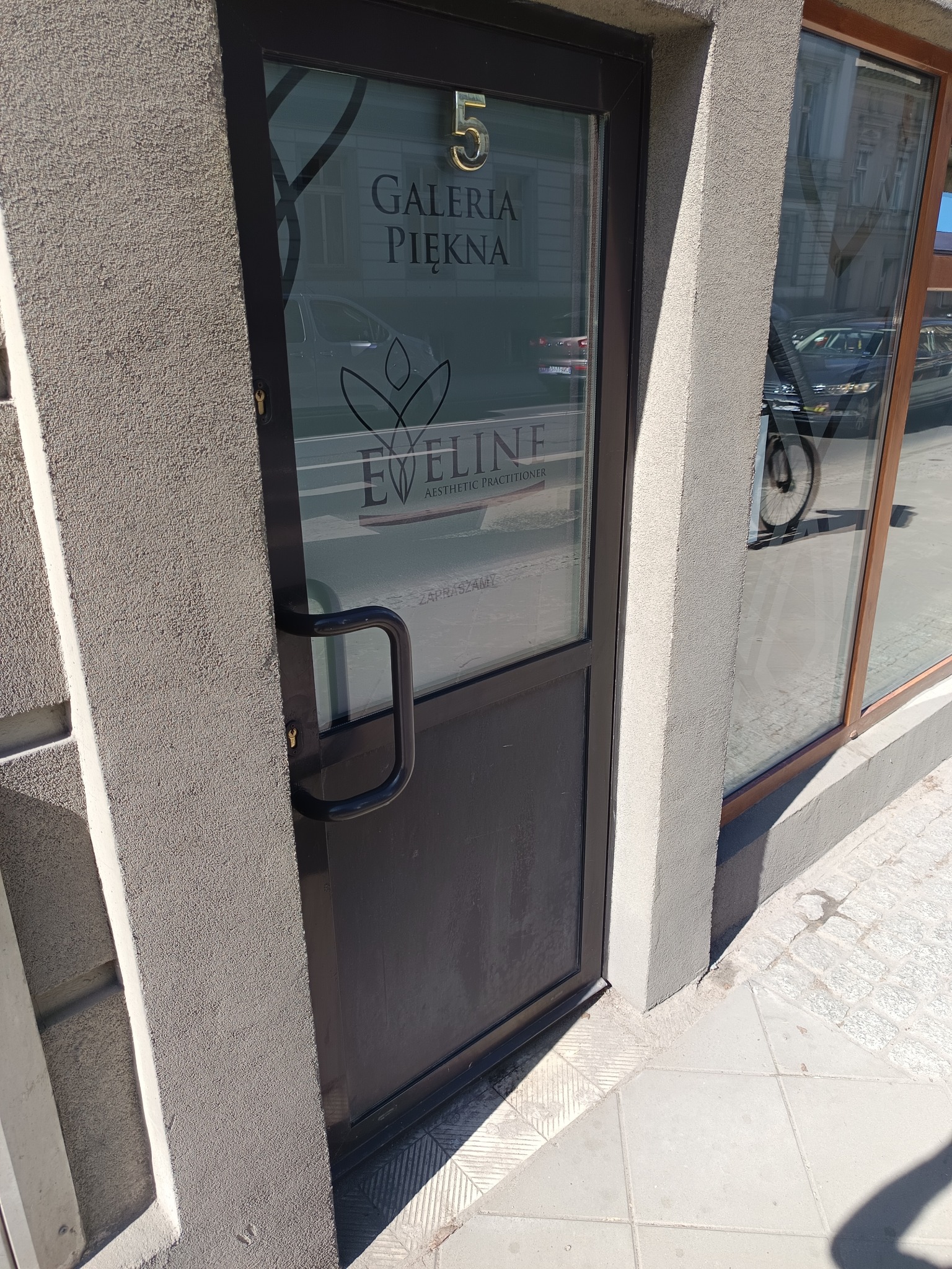 Szklane drzwi wejściowe do 'Galerii Piękna' z numerem 5 nad napisem. Widoczne odbicie ulicy i fragment okna sąsiedniego lokalu.