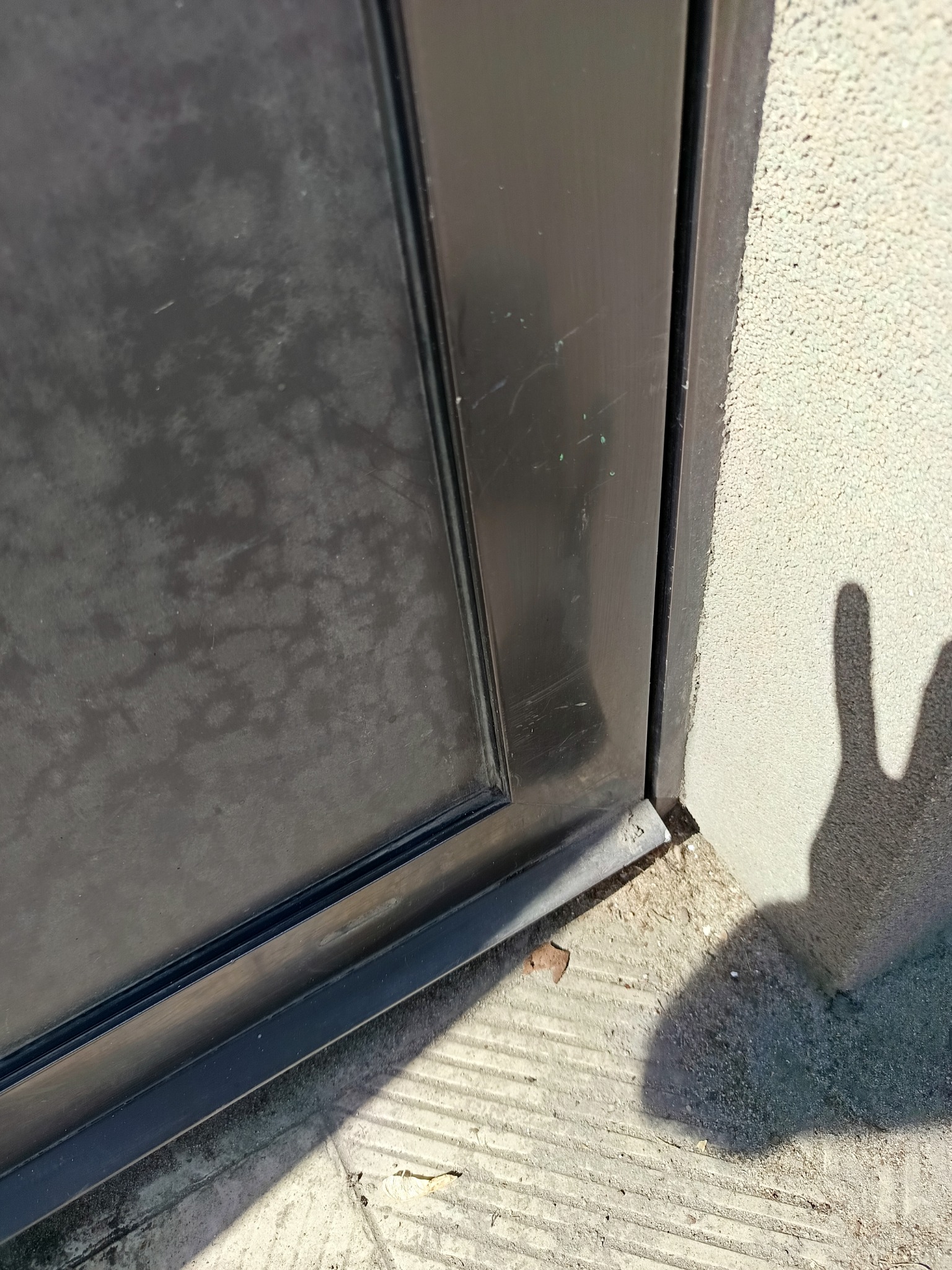 Zabrudzone, czarne okno z widocznym cieniem dłoni na ścianie. Widoczne ślady po zaciekach i zabrudzeniach. Narożnik okna i fragment ściany z fakturą.