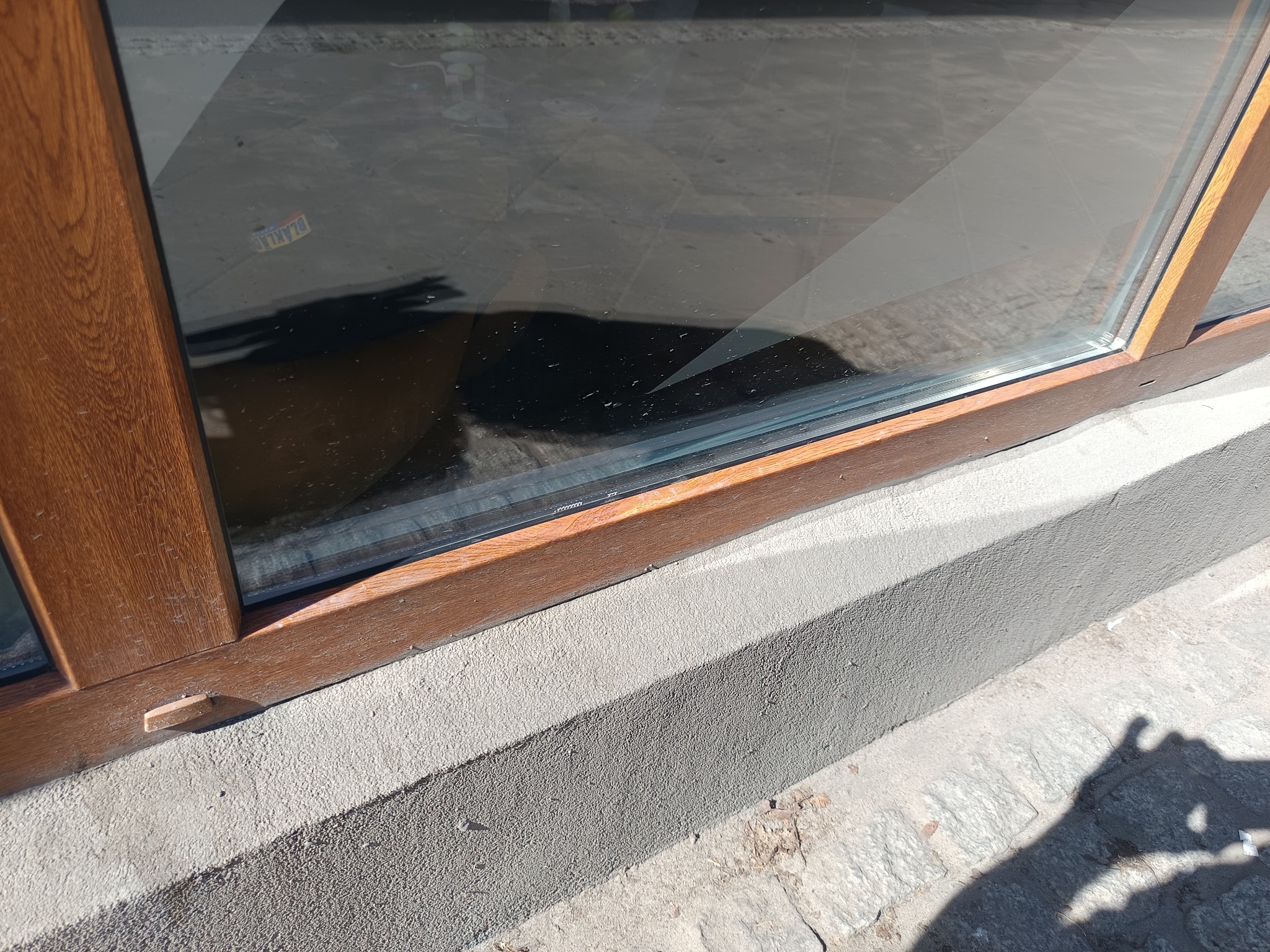 Czysta szyba okienna z drewnianą ramą, widoczny fragment elewacji i chodnika. Na szybie widoczna naklejka. Cień osoby fotografującej.