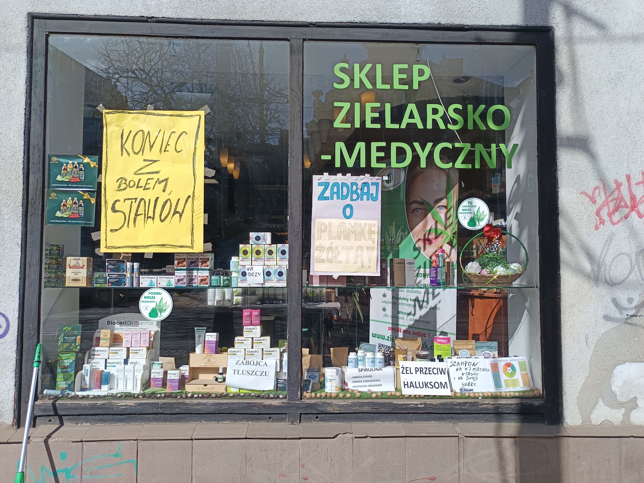 Witryna sklepu zielarsko-medycznego z plakatami informacyjnymi i produktami na półkach. Widoczny napis 'Sklep Zielarsko-Medyczny' i hasła reklamowe.