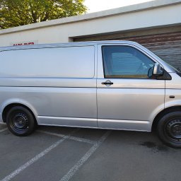 Piotr Kukula - Srebrny bus dostawczy zaparkowany na parkingu, widok z boku, z czarnymi felgami i oponami. W tle budynek z szarą bramą garażową i zielonym drzewem.