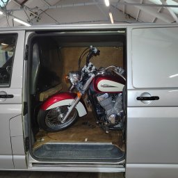 Piotr Kukula - Czerwono-biały motocykl w srebrnym busie z otwartymi drzwiami przesuwnymi. Wnętrze busa wyłożone sklejką. Widoczna część dachu z oświetleniem.