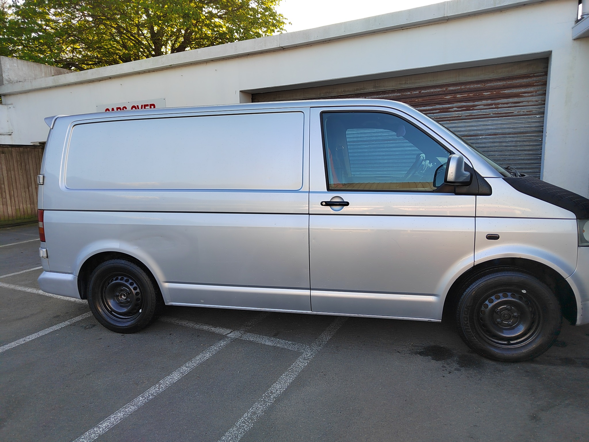 Srebrny bus dostawczy zaparkowany na parkingu, widok z boku, z czarnymi felgami i oponami. W tle budynek z szarą bramą garażową i zielonym drzewem.