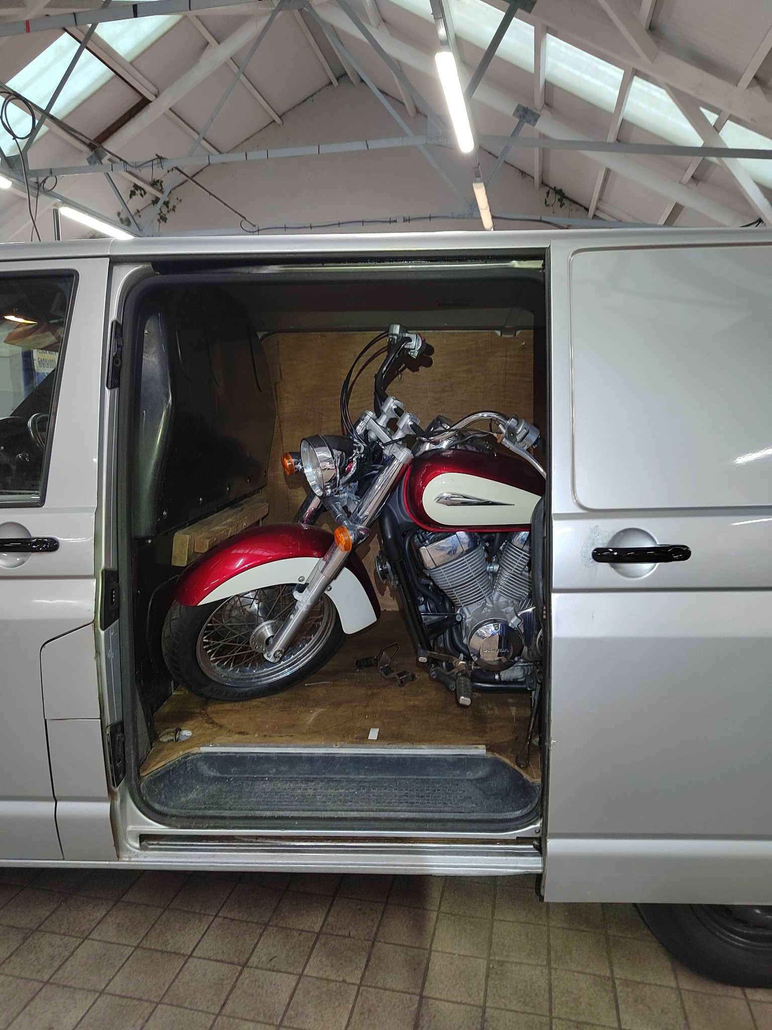 Czerwono-biały motocykl w srebrnym busie z otwartymi drzwiami przesuwnymi. Wnętrze busa wyłożone sklejką. Widoczna część dachu z oświetleniem.