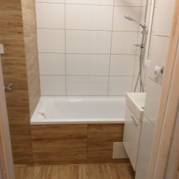 Mixbud - Nowoczesna łazienka z wanną obudowaną drewnopodobnymi płytkami, białą armaturą i kabiną prysznicową. Podłoga wyłożona panelami imitującymi drewno.