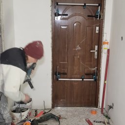 Mixbud - Montaż brązowych drzwi wewnętrznych z użyciem rozpórek. Widoczne narzędzia, poziomica i odkurzacz. Fachowiec w trakcie pracy, w tle białe ściany i płytki podłogowe.