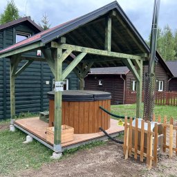 Ibiza - Balie & Sauny - Zadaszona, drewniana balia na tarasie z drewnianą obudową i piecem na zewnątrz. W tle domki z ciemnego drewna i zieleń. Całość w naturalnym otoczeniu.