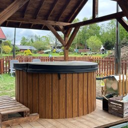 Ibiza - Balie & Sauny - Okrągła, drewniana sauna zewnętrzna pod zadaszeniem, z paleniskiem i schodkami, na tarasie kompozytowym. W tle zielony ogród i zabudowa jednorodzinna.