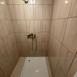 PUDOX - Wykończony brodzik z beżowymi płytkami imitującymi drewno. Widoczna bateria prysznicowa i otwór po słuchawce. Minimalistyczny design kabiny.