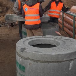 FIRMA BUDOWLANA MARITIME - Montaż betonowych kręgów studni przez robotników w pomarańczowych kamizelkach. Widoczne rury kanalizacyjne i sprzęt budowlany w tle. Praca w toku.