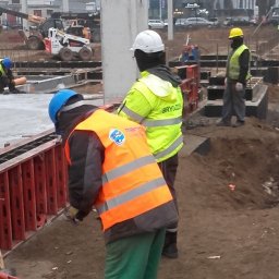 FIRMA BUDOWLANA MARITIME - Praca wre na budowie: robotnicy w kaskach i kamizelkach montują szalunki fundamentów. W tle widoczny sprzęt budowlany i betonowe elementy konstrukcyjne.