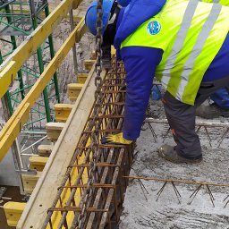 FIRMA BUDOWLANA MARITIME - Pracownik w kasku i kamizelce układa zbrojenie w szalunku fundamentu. Widoczne żółte elementy szalunku i wystające pręty zbrojeniowe z betonu.