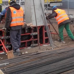 FIRMA BUDOWLANA MARITIME - Pracownicy w odblaskowych kamizelkach przy szalunkach fundamentowych. W tle widoczne zbrojenia i sprzęt budowlany. Prace ziemne w toku.