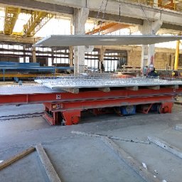 FIRMA BUDOWLANA MARITIME - Płyta prefabrykowana zbrojona siatką stalową, podnoszona w hali produkcyjnej za pomocą suwnicy. Widoczny wózek transportowy i elementy konstrukcyjne budynku.