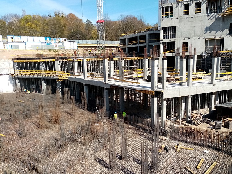 Wysoki budynek w trakcie budowy, widoczne zbrojenia i betonowe kolumny, na górze pracownicy w żółtych kamizelkach, w tle drzewa i niebo.