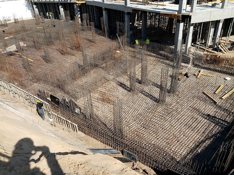 Zbrojenie fundamentów pod budynek wielopiętrowy, widok z góry na siatkę zbrojeniową i wystające pręty. W tle widoczni pracownicy w odblaskowych kamizelkach.