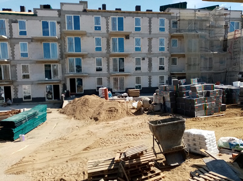 Wznoszony budynek wielorodzinny z betonu komórkowego. Widoczne okna, rusztowania, materiały budowlane i teren budowy z piaskiem. Mieszarka do betonu w tle.