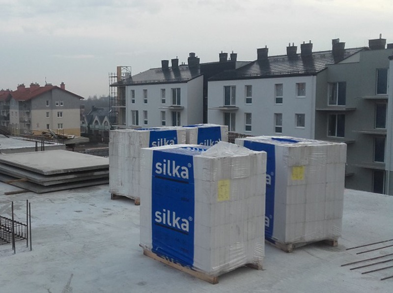 Cegły Silka na paletach na budowie wielorodzinnej, widok z góry. W tle budynki w trakcie budowy, szare niebo. Elementy zbrojenia na pierwszym planie.