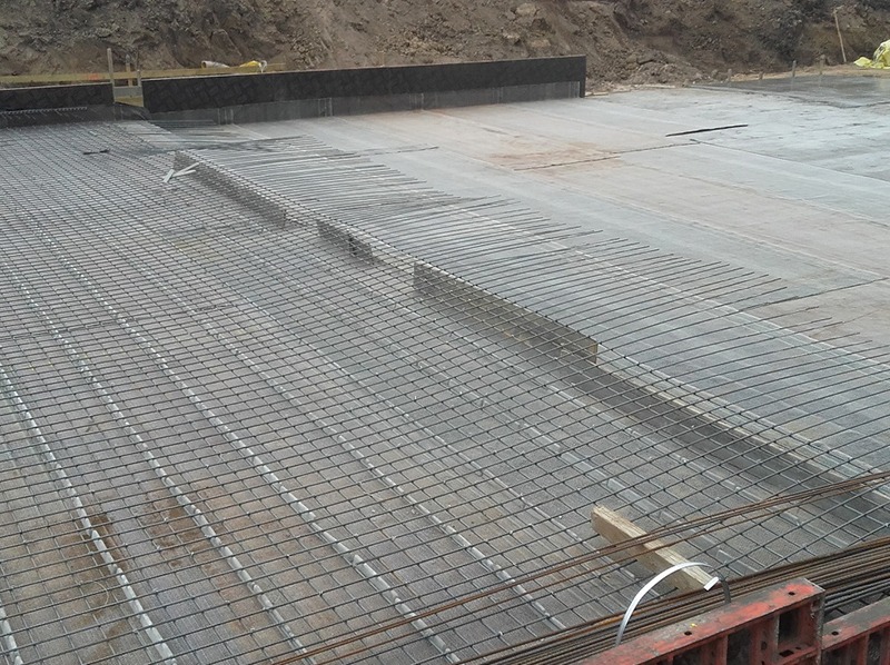 Zbrojenie płyty fundamentowej z siatki stalowej na folii, przygotowane do zalania betonem. Widoczne elementy szalunku i ziemia w tle.