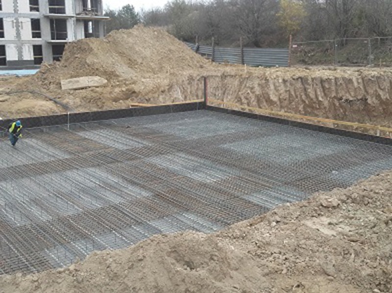 Zbrojenie fundamentu pod budynek wielorodzinny, widoczne siatki stalowe i wykop. W tle hałda ziemi i budynek w trakcie budowy. Pracownik w kasku przy zbrojeniu.