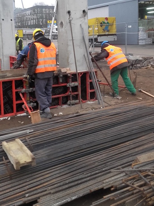 Prace zbrojarskie na budowie: robotnicy w kamizelkach układają stalowe pręty w szalunkach pod betonowe słupy. Na pierwszym planie wiązki prętów zbrojeniowych.