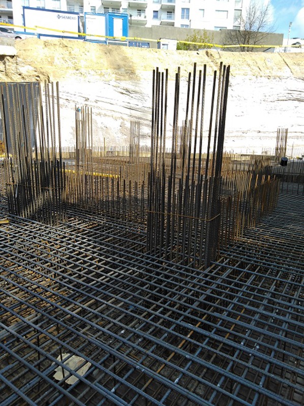 Zbrojenie fundamentów z pionowymi prętami na placu budowy, widok z góry na siatkę zbrojeniową. W tle ziemny nasyp i budynek mieszkalny.