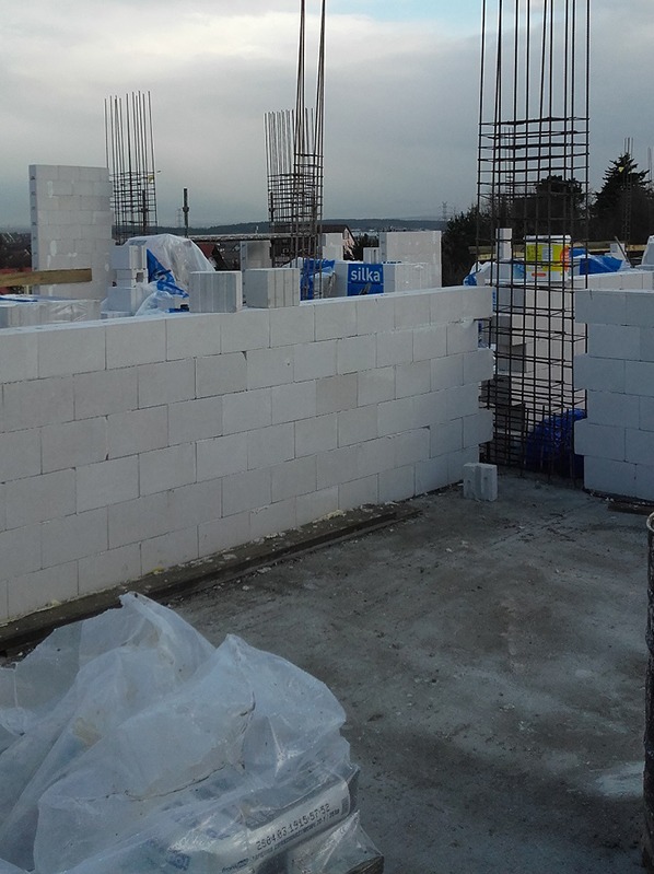 Wznoszenie muru z białych bloczków Silka, widoczne zbrojenia pionowe. Częściowo zakryte materiały budowlane w folii. Budowa w toku, pochmurne niebo w tle.