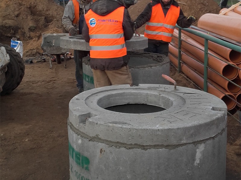 Montaż betonowych kręgów studni przez robotników w pomarańczowych kamizelkach. Widoczne rury kanalizacyjne i sprzęt budowlany w tle. Praca w toku.