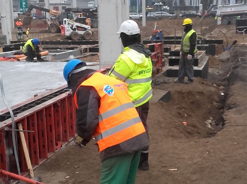 Praca wre na budowie: robotnicy w kaskach i kamizelkach montują szalunki fundamentów. W tle widoczny sprzęt budowlany i betonowe elementy konstrukcyjne.