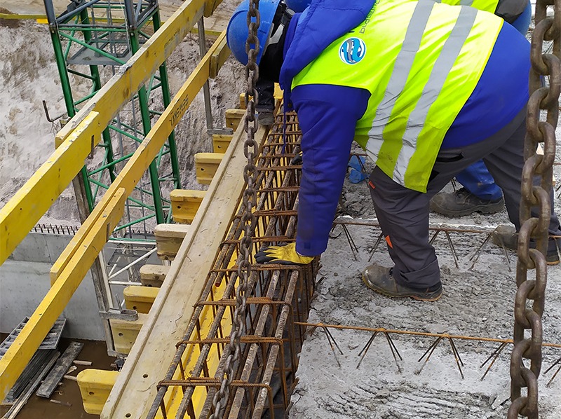Pracownik w kasku i kamizelce układa zbrojenie w szalunku fundamentu. Widoczne żółte elementy szalunku i wystające pręty zbrojeniowe z betonu.