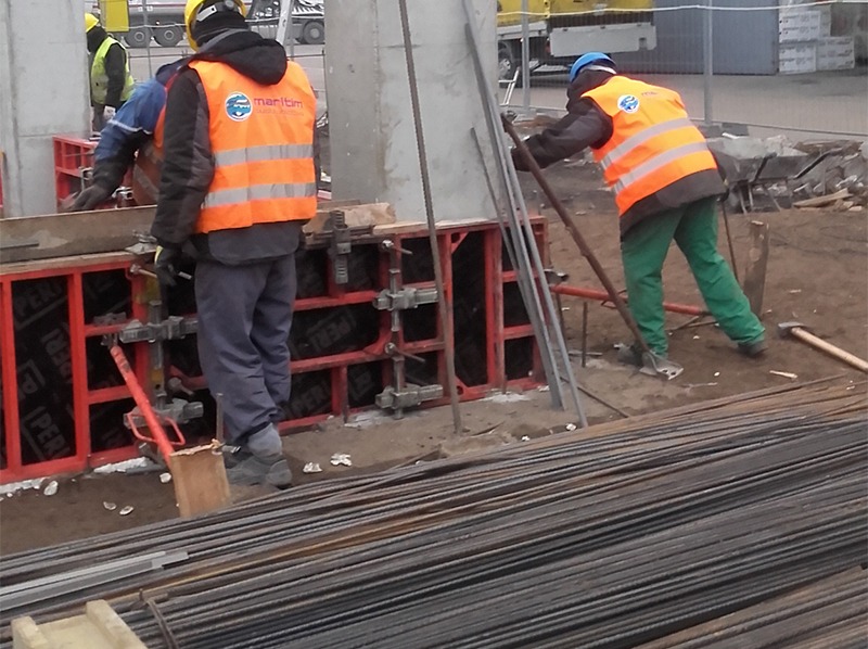 Pracownicy w odblaskowych kamizelkach przy szalunkach fundamentowych. W tle widoczne zbrojenia i sprzęt budowlany. Prace ziemne w toku.