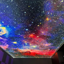 USŁUGI REMONTOWO-WYKOŃCZENIOWE DANIEL TĘCZA - Sufit napinany z motywem kosmosu, widok z dołu. Efektowna instalacja w pomieszczeniu z panelami akustycznymi. Intensywne kolory i detale gwiazd.
